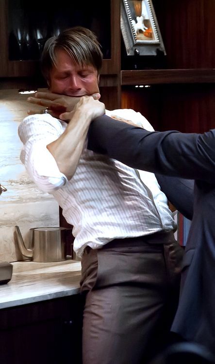 mads mikkelsen being strangled in a kitchen. he is wearing a white and light blue striped shirt and brown pants. a man in a navy blue suit is strangling him. they are standing by a counter in a kitchen.