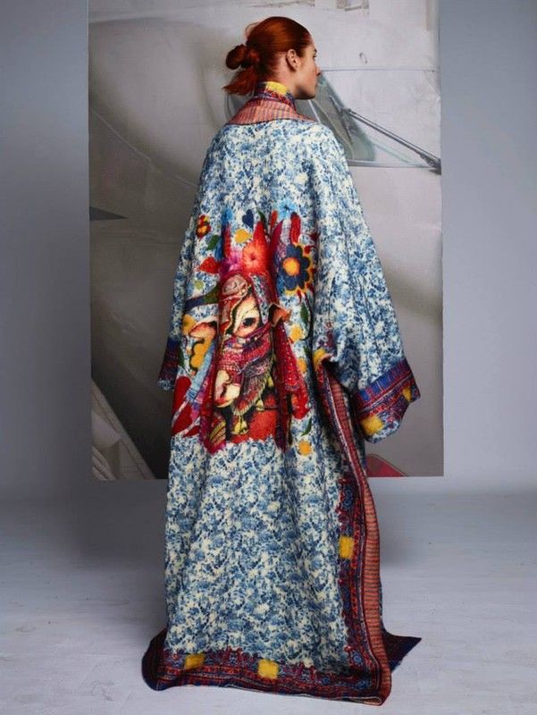 the image shows a woman from the back wearing a long, loosefitting coat. the coat is predominantly offwhite or beige with a blue and white patterned base.  a large, vibrant, handpainted illustration dominates the back of the coat, depicting a scene with figures, flowers, and other elements in rich, bold colors. the coat has a loose, flowing shape and what looks to be a high collar or neck. the model has red hair tied back in a bun. the backdrop is simple and muted, creating contrast with the coat's intricate design. the overall aesthetic suggests a highfashion or artistic clothing piece.