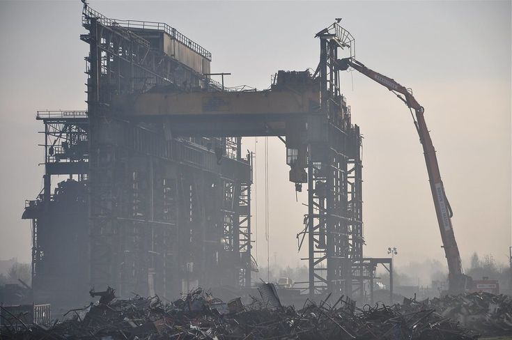 the image shows the demolition of a large industrial structure, possibly a factory or steel plant. a massive crane is actively removing parts of the building, while the foreground is filled with a large pile of scrap metal and debris from the demolition process.  a haze or light smoke hangs in the air, obscuring the background slightly. the overall scene is one of largescale industrial dismantling in somewhat gloomy atmospheric conditions.