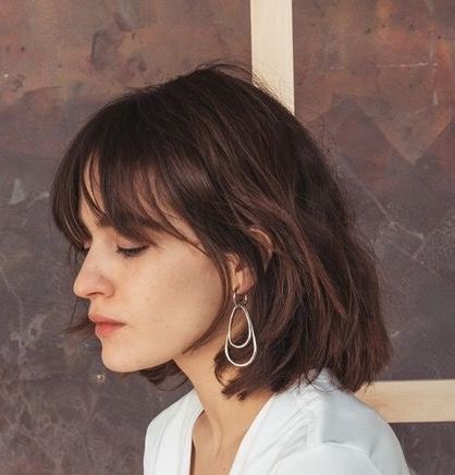 a profile portrait of a woman with a bob haircut and bangs, wearing teardrop earrings and a white shirt, against a blurred background.