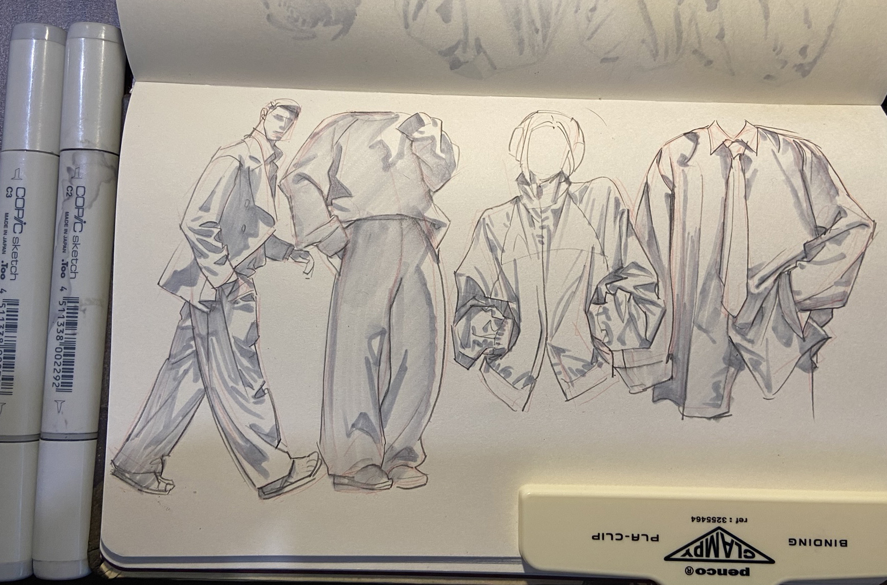 a sketchbook page with four fashion illustrations of men's clothing drawn with gray copic markers. the illustrations depict oversized jackets and baggy pants in a contemporary style. two copic markers and a clipboard are laying on top of the page.
