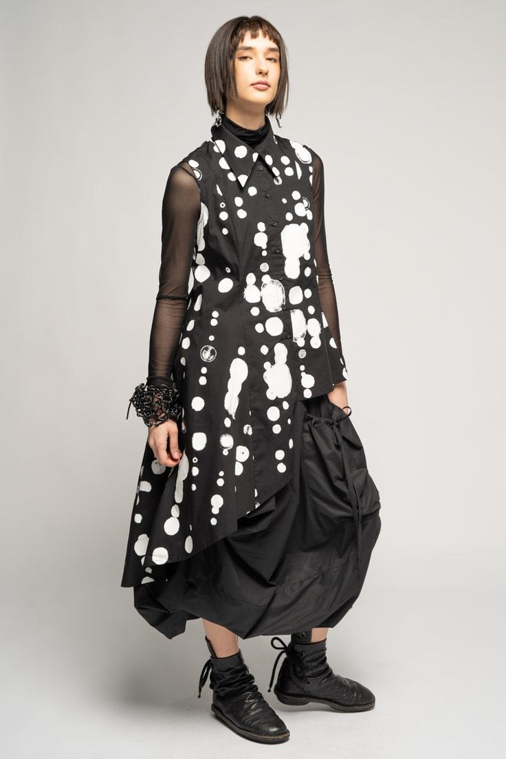 a young woman with short dark hair is wearing a black and white polka dot dress with a black skirt. she is wearing black boots and standing on a grey background.