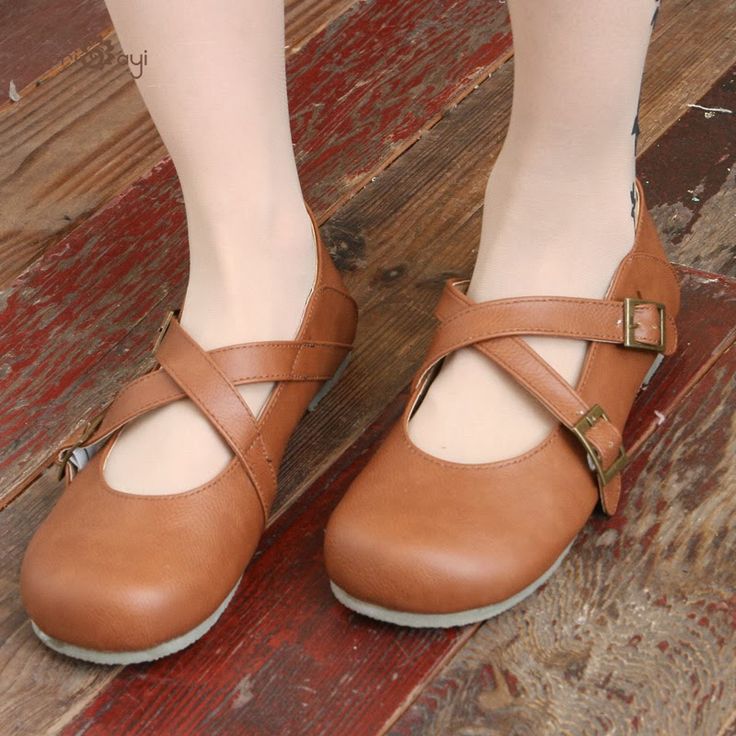 a closeup shot shows a pair of brown, flat shoes with a cross strap design and a buckle on the side. the shoes are being worn, and the photo is taken indoors, with a wooden floor as the background. the focus is on the shoes and their details.