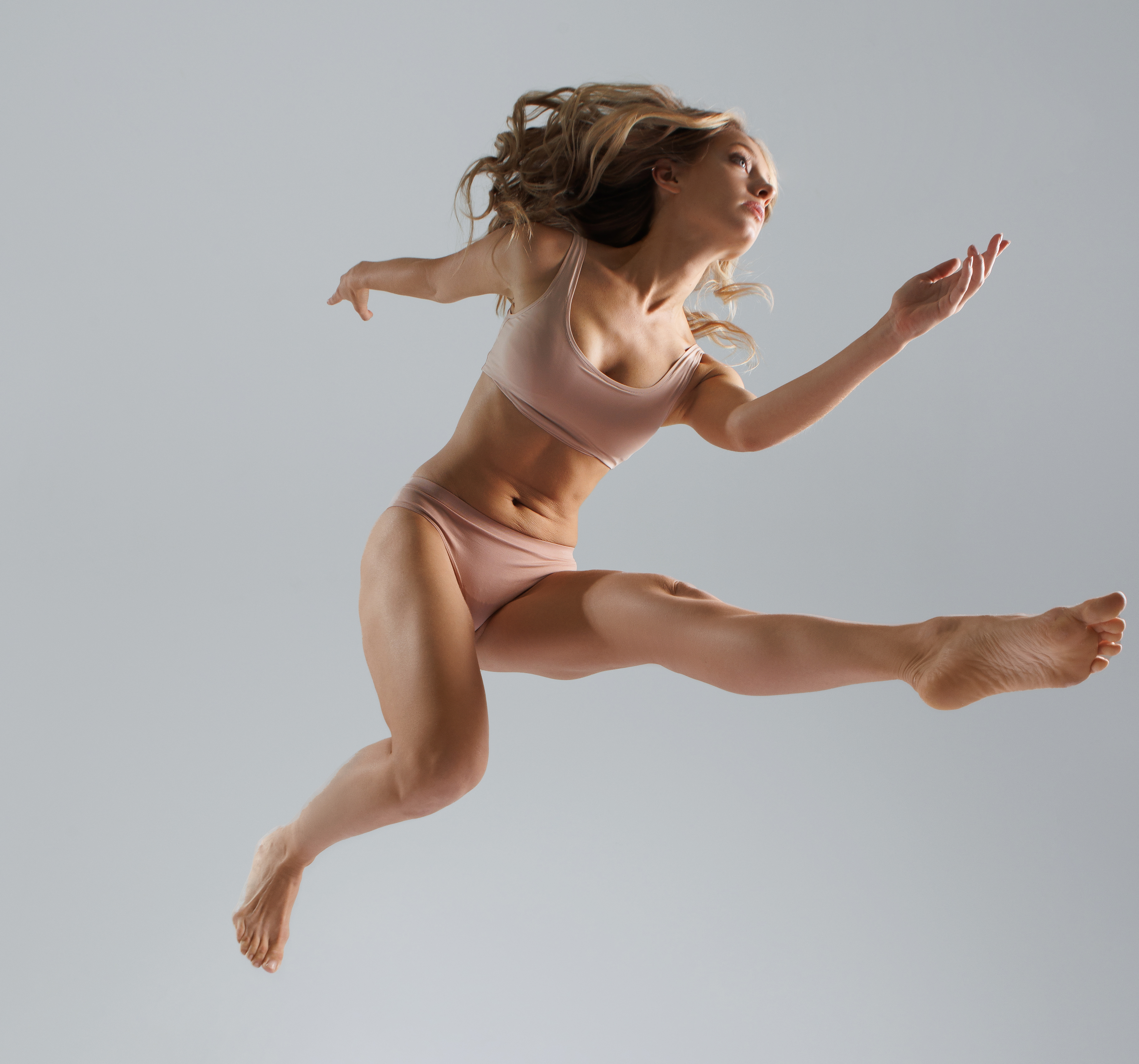 a woman with blonde hair is captured midair in a dynamic pose. she is dressed in a nudecolored sports bra and matching underwear, emphasizing her athleticism. her body is toned and muscular, showcasing her dedication to exercise. she is barefoot, suggesting a connection to the earth and freedom of movement. the background is a plain gray, which makes her actions pop. her body is stretched out in a horizontal jump, suggesting effort, skill, and power.