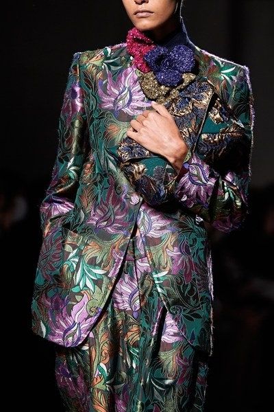 the image showcases a model on a runway, wearing a striking pantsuit. the suit is a rich brocade fabric featuring an elaborate, jeweltoned floral pattern in shades of deep green, purple, and gold.  the pattern is highly detailed and appears to incorporate embroidery or similar techniques. the model is wearing a large, ornate brooch or collar composed of different textured fabrics and jewels at the neckline.  the dark background highlights the richness of the clothing's colors and texture. the overall look is sophisticated, luxurious, and indicative of highend fashion. the image's focus is on the intricate details of the suit's design and the model's elegant presentation.