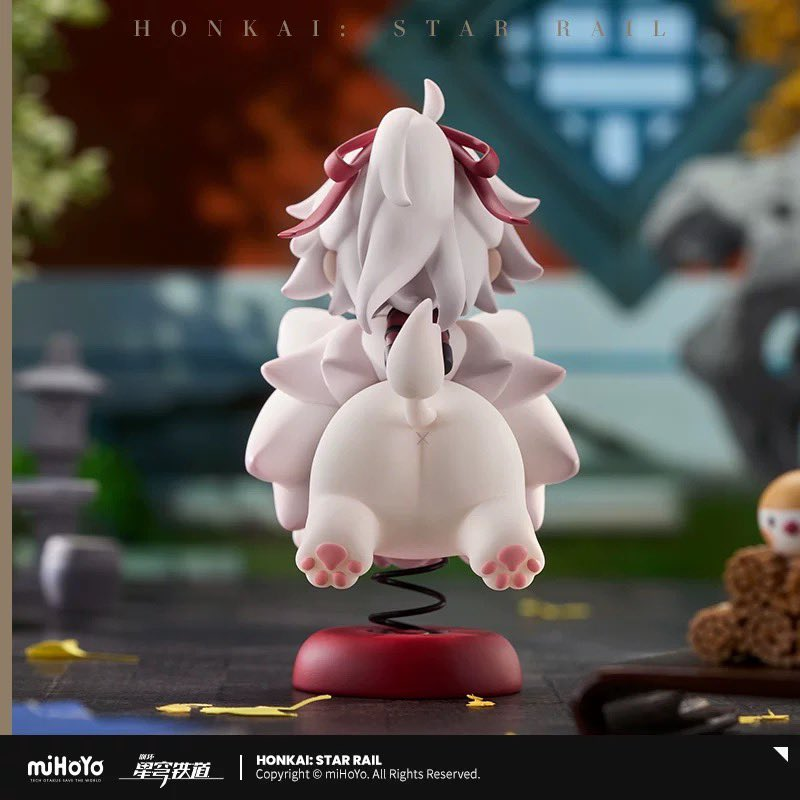 a cute anime character figurine from the game "honkai: star rail" by mihoyo.  the figurine is small, white, and sitting on a red spring. the character is wearing a white and red outfit, and has a red ribbon on their head. the background of the image is blurred and out of focus.