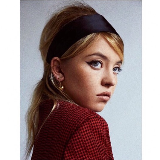 the image is a closeup portrait of a young woman with a stylish 1960s inspired beehive hairstyle.  she is wearing a black satin headband, a red houndstooth jacket, and gold earrings. her makeup includes dramatic winged eyeliner. the background is a plain light grey, drawing focus to the woman's face and attire.  the overall style is retrochic and elegant.