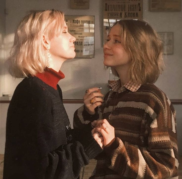 two young women are standing close to each other, holding hands and smiling as they look into each other's eyes. the girl on the left is wearing a black sweater and a red scarf while the girl on the right is wearing a brown sweater with a plaid shirt underneath. they are both inside a building, and it appears to be a sunny day. they are both giving off a soft, grunge aesthetic. the image is likely taken from the waist up.
