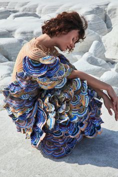 a young woman with curly brown hair, wearing a colorful ruffled dress, sits on a white background. the dress has multiple layers and has  blue, yellow, and brown hues. she is looking down, posing for a fashion portrait, with her hands gently touching the ground. the background is made of white textured surface.