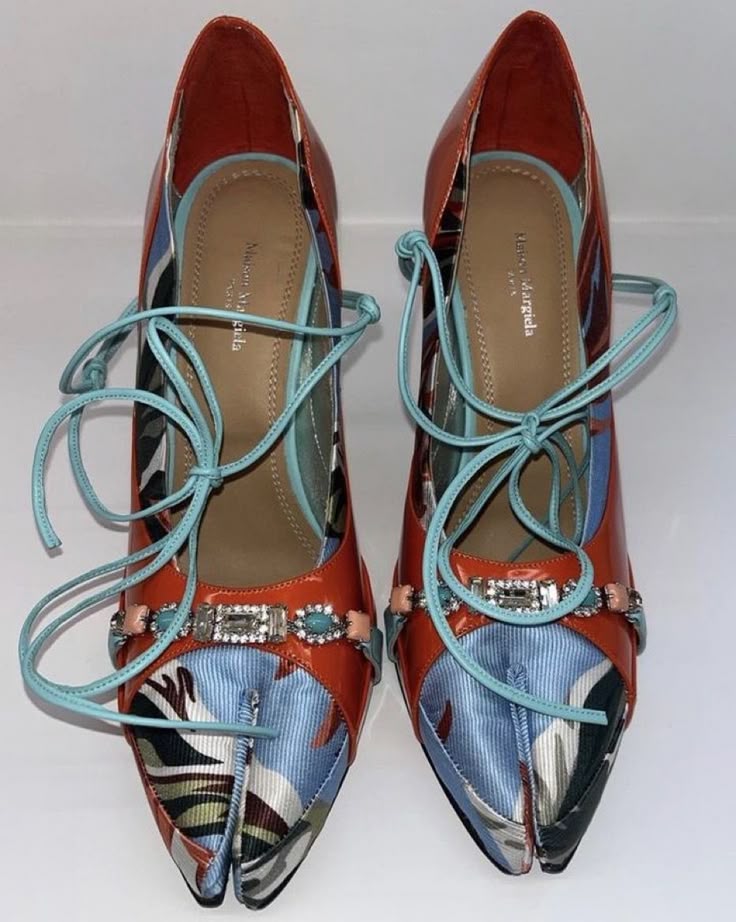 two orange and blue patterned dress shoes with laces are displayed in front of a white background. the shoes have a splittoe design and decorative embellishments on the straps.