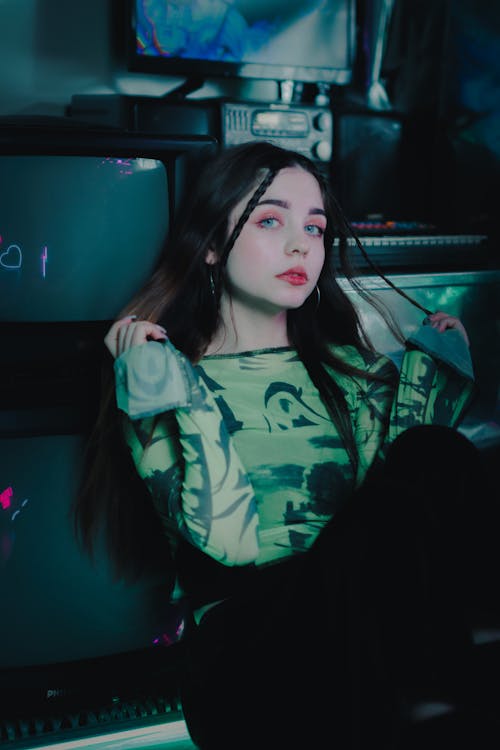 a young woman with long dark hair sits in front of several computer screens and other vintage electronics. she's wearing a green mesh longsleeved shirt and dark pants. the image has a moody, retro aesthetic with dark lighting and vibrant colors. she is looking directly at the camera, with pink eyeshadow adding to the stylistic look.  the background is cluttered with older technology.
