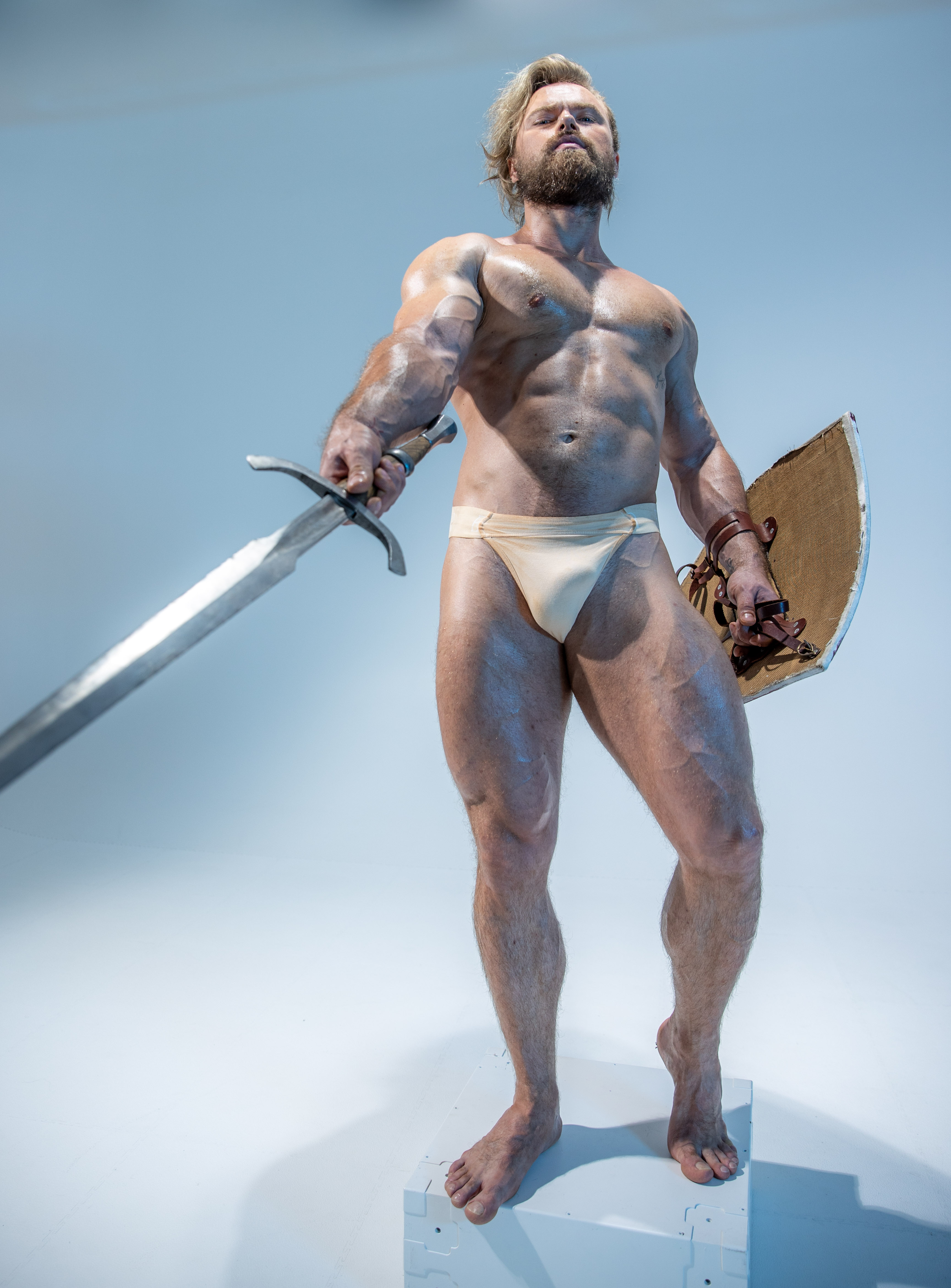 a muscular man, oiled and glistening, stands on a white pedestal against a pale blue background. he adopts a warrior's stance, holding a gleaming sword in his right hand and a round shield in his left. his attire is minimal, consisting only of a lightcolored loincloth.  the image evokes a sense of strength, power, and perhaps a connection to ancient warriors or mythological figures.