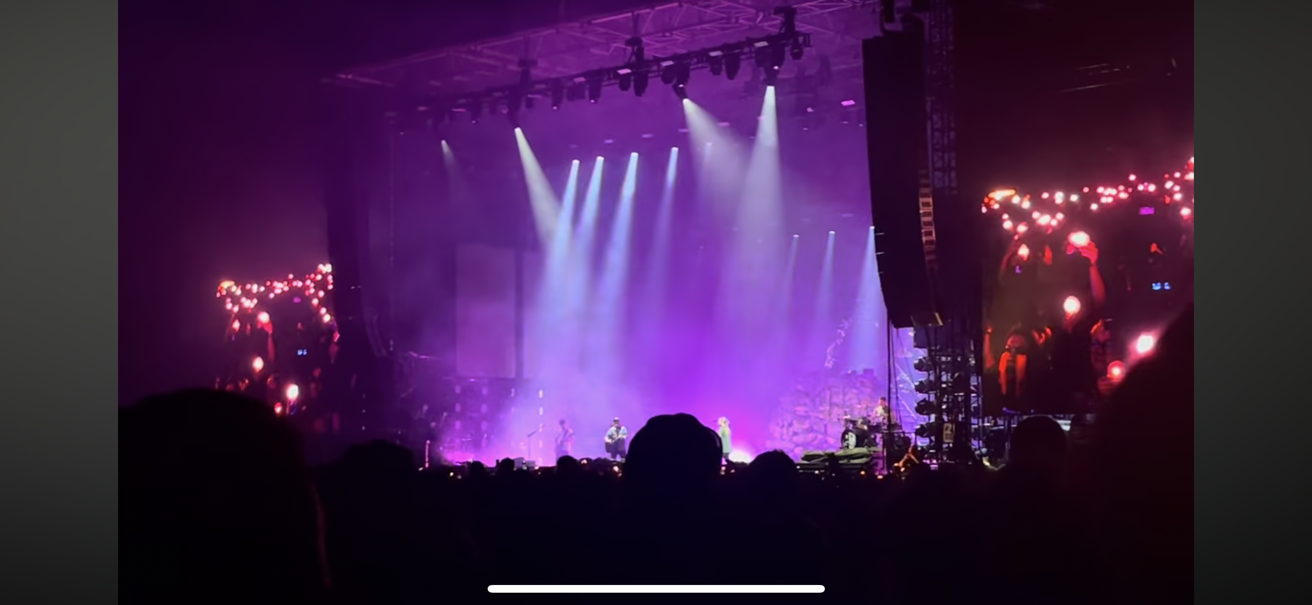 this image captures a vibrant concert scene, with a band performing on a stage illuminated by dazzling lights. the stage is shrouded in a purple haze, creating an ethereal atmosphere. the crowd, silhouetted in the darkness, waves their hands in the air, reveling in the music and energy of the performance.