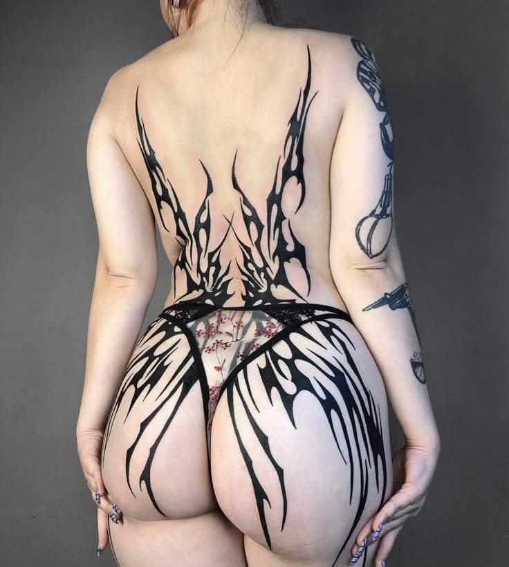 a woman is photographed from the rear, showcasing a large tribalstyle tattoo that extends from her upper back down to her buttocks. the woman is wearing floral lingerie that is partially obscured by the tattoo. additional arm tattoos are visible on her left arm. her right hand is visible in the bottom left corner of the image. she is against a plain grey background.