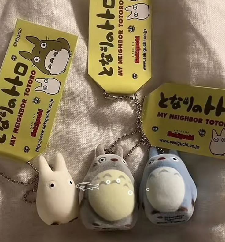 the image shows three totoro keychains with their accompanying tags. the keychains are small plush toys representing different characters from the animated film "my neighbor totoro." the characters represented are a white mini totoro, a grey totoro with a yellow belly, and a blue totoro. each keychain is attached to a yellow tag that features the title of the film in japanese and english, the sekiguchi brand name, and the website address. the overall tone is whimsical and nostalgic, appealing to fans of the film.