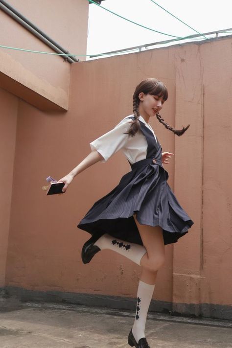 a young woman in a white shirt and navy blue pinafore dress is captured middance on a rooftop.  she's wearing white socks with black accents and black mary jane style shoes. her long dark hair is styled in two braids. she holds a small notebook in her right hand. the background features a peachcolored building with visible pipes and wires. the image is taken from a low angle, emphasizing her movement and the urban environment. the lighting is natural and overcast, creating a soft and diffused effect.