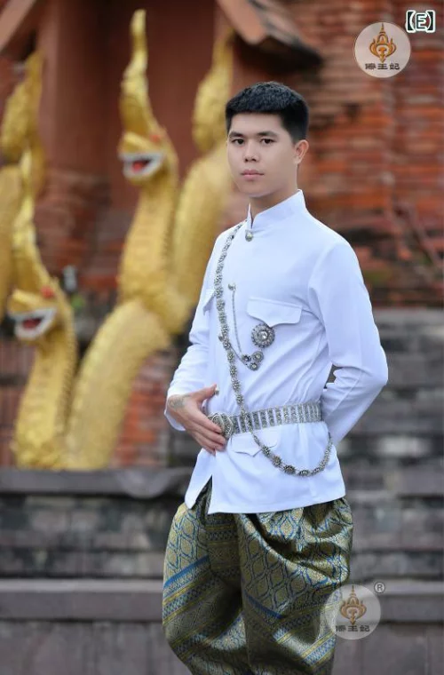 a man is standing in front of a temple wearing a traditional white thai outfit. the man is wearing a white shirt with a gold trim and a patterned gold and silver belt. he is also wearing a patterned gold and silver fabric around his legs. the man is smiling and looking at the camera. the man is standing in front of a temple, which is decorated with gold and silver details.