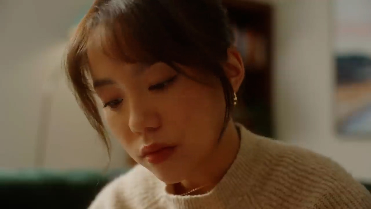 a young woman sits indoors, her head tilted slightly down in thought. the warm, artificial lighting illuminates her face, highlighting her features. she is wearing a cozy, beige sweater, suggesting a comfortable and intimate atmosphere.  the focus on her contemplative expression invites the viewer to wonder about her thoughts.