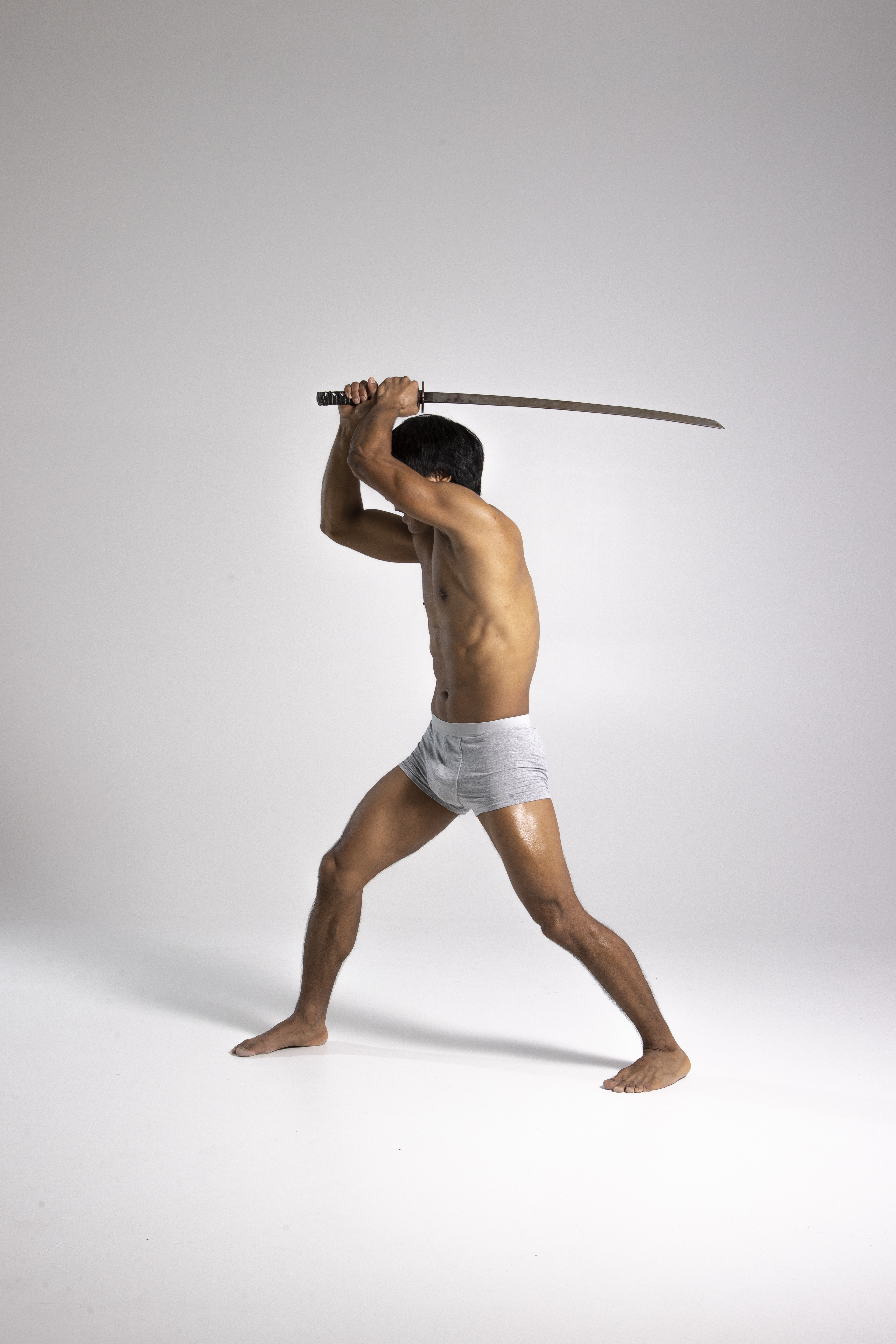 a shirtless asian man in gray underwear stands barefoot against a white background, holding a katana sword above his head in a martial arts pose. he appears strong and determined, ready for battle.