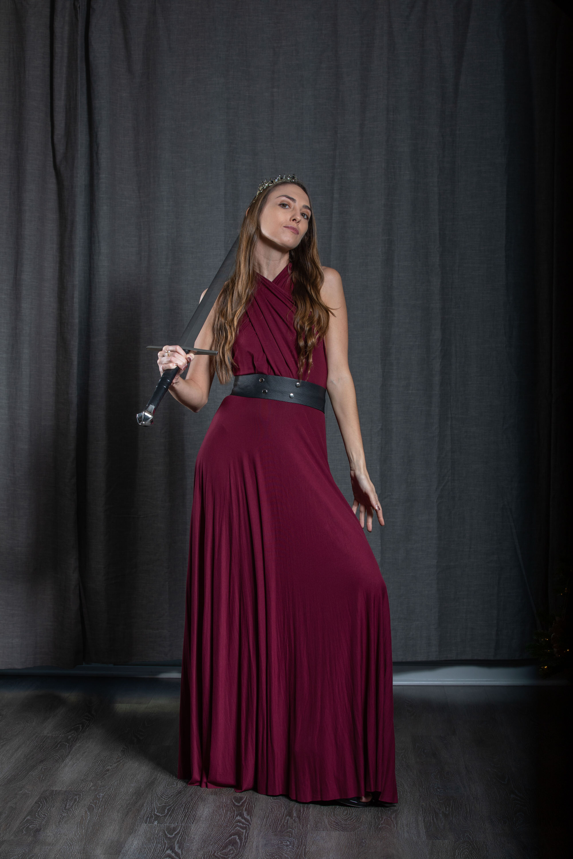 a young woman stands gracefully in a flowing maroon dress, holding a sword casually at her side.  she wears a delicate crown upon her head, conveying an air of regality and strength.  she poses before a dark gray curtain backdrop in a studio setting, bathed in soft, artificial lighting.