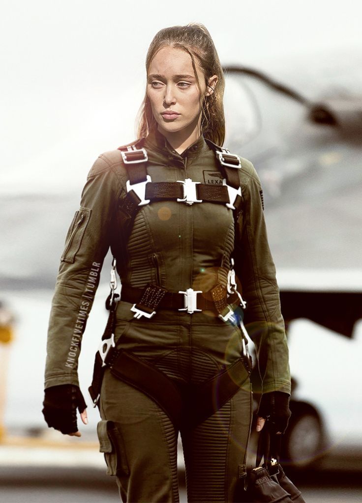 a woman in a green flight suit and black gloves is standing on an airport runway with an airplane in the background. she is looking away from the camera with a serious expression on her face. she is wearing a harness and a belt. she has long, dark hair pulled back in a ponytail.  the sun is shining brightly in the background.  the woman is giving off a very intense and determined vibe.