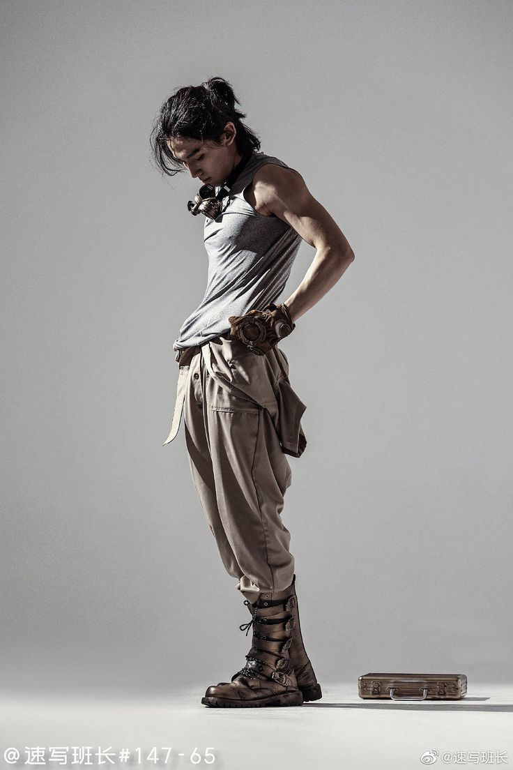 a full body studio shot of a man wearing a steampunkinspired outfit. the man is leaning forward, looking down. he has a tank top, tan pants, boots, gloves, and goggles around his neck. a metal briefcase sits on the floor nearby. the background is a grey gradient.