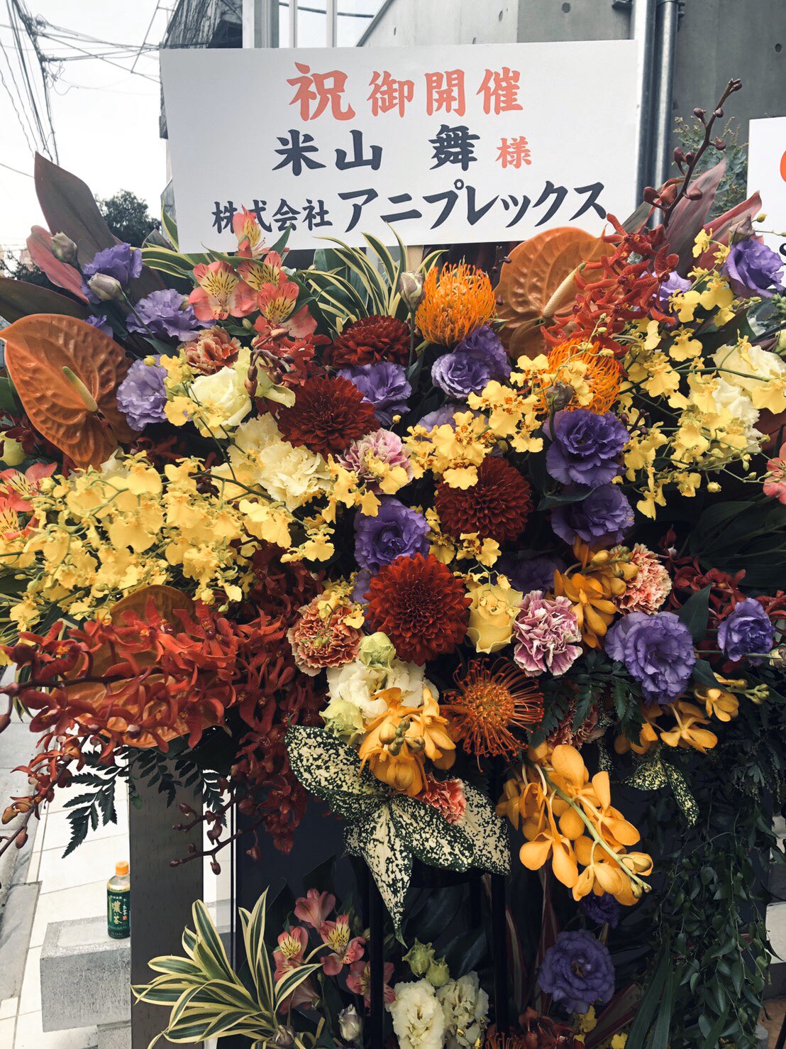 a vibrant flower arrangement with red, orange, yellow, and purple flowers stands outside. a sign with japanese text celebrating a grand opening is attached to the arrangement.