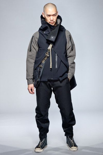 a man is standing in a studio setting, wearing a layered outfit consisting of a gray hooded jacket, a black vest with multiple pockets, and black pants. he is also wearing black and white sneakers. his head is shaved, and he has a serious expression on his face. the image is welllit and shows off the details of the clothing.