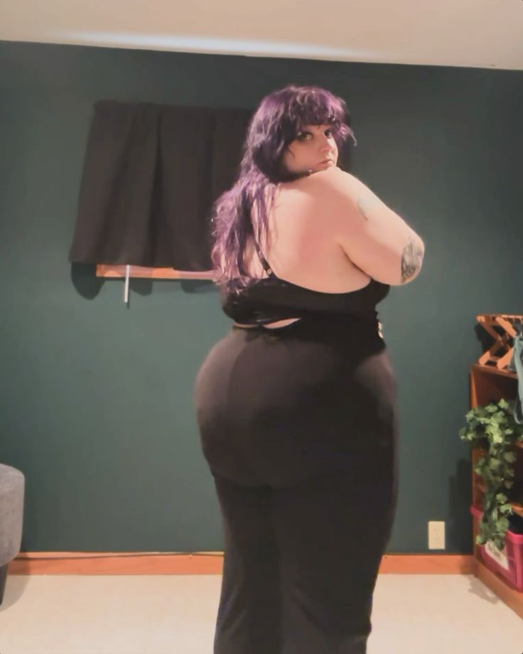 a plus size woman with purple hair is posing in a black outfit. she is looking over her shoulder at the camera. the background is a dark green color. she is standing indoors. the woman has a curvy figure and a tattoo on her arm.