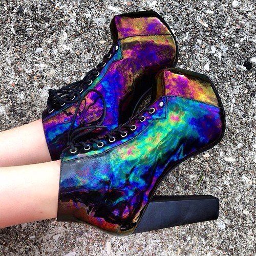 a pair of iridescent, laced highheeled boots sit on a rough, grey surface. the boots have a distinctive holographic effect with a multitude of colors, and blocky, rectangular heels. the laces are black. only the wearer's lower legs and feet in the boots are visible.