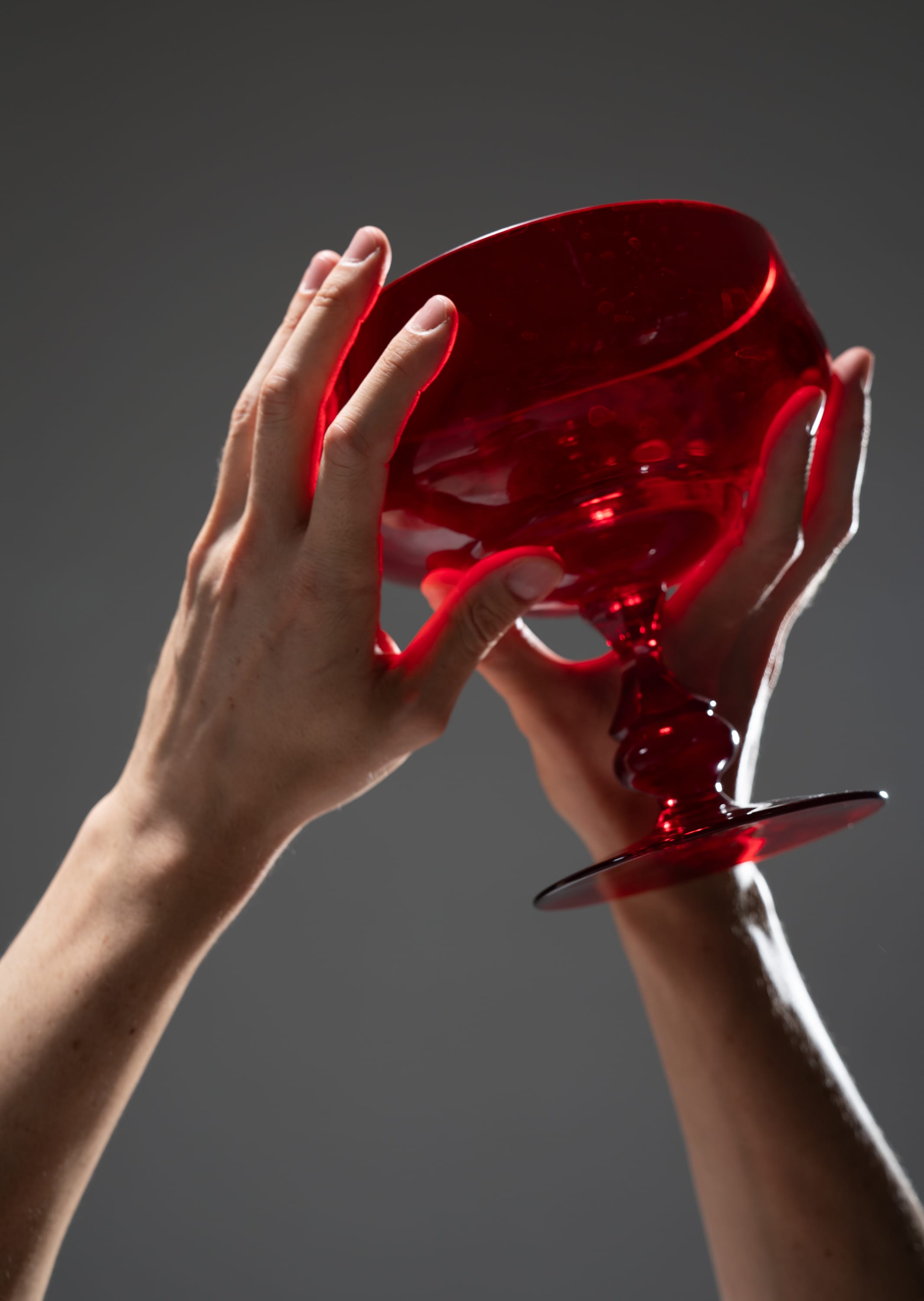 a pair of hands gently lift a brilliant crimson goblet towards a dimly lit background. the light pierces through the glass, illuminating the hand and casting a warm, reddish glow. the perspective from below emphasizes the gesture of raising the goblet, creating a sense of anticipation or celebration.