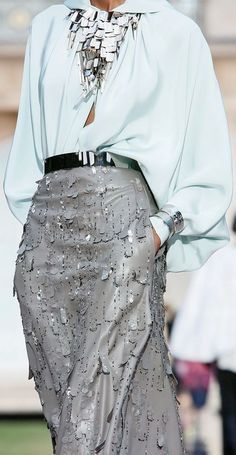 a woman is standing on a street, wearing a silver sequined skirt, a white blouse with a silver necklace and a black belt. she is posing with a serious face and looking to the left side. the background is blurred and has people standing in the background.