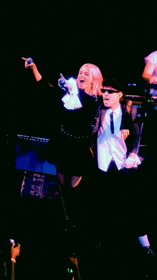 two performers, a woman and a man, are on stage singing into microphones. the woman has blonde hair and is wearing a black polka dot dress with white ruffled sleeves. she is pointing with one hand. the man is wearing sunglasses and a black suit with a striped jacket. he has a white shirt and a black tie. they are performing in front of a stage with musical equipment.