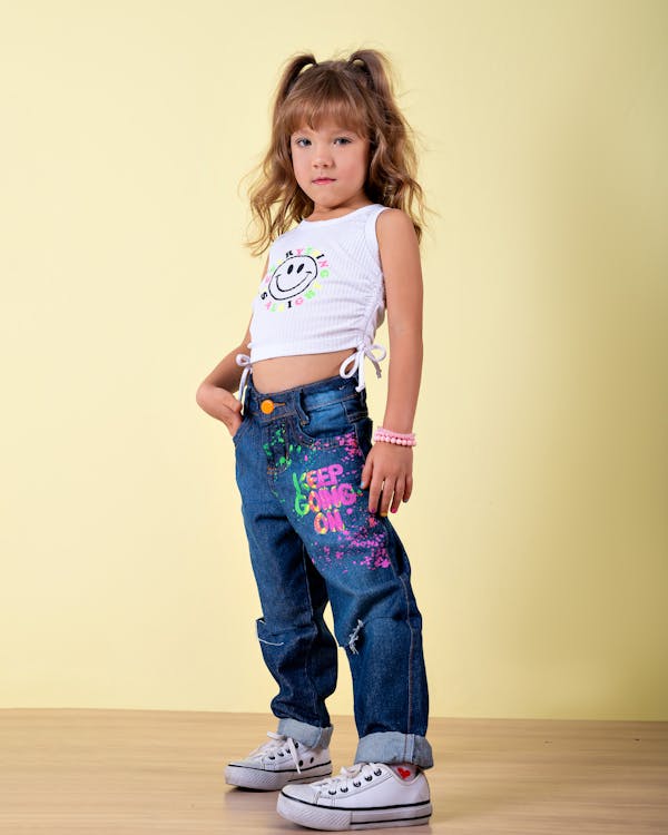 a fulllength portrait of a young girl standing in a studio against a pale yellow backdrop. she is wearing a white ribbed tank top with a smiley face design, denim jeans with colorful graffitistyle writing and paint splatters, and white converse sneakers. her hair is styled in pigtails with bangs, and she is wearing a pink bracelet. she is posing with one hand on her hip, looking directly at the camera.