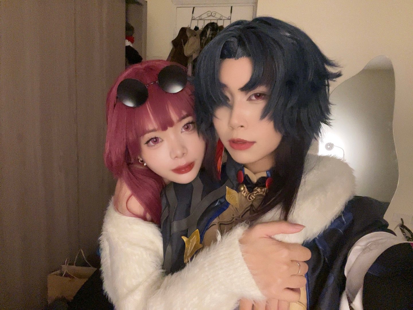 a closeup portrait of a couple cosplaying anime characters. the girl is wearing a white fur coat and has long red hair and a black headband. the boy is wearing a blue and black outfit with a white fur collar and has blue hair. they are both smiling and looking at the camera. they are hugging, and the girl has her arms around the boy's waist. they appear to be happy and having fun. the image is welllit and has a warm tone.