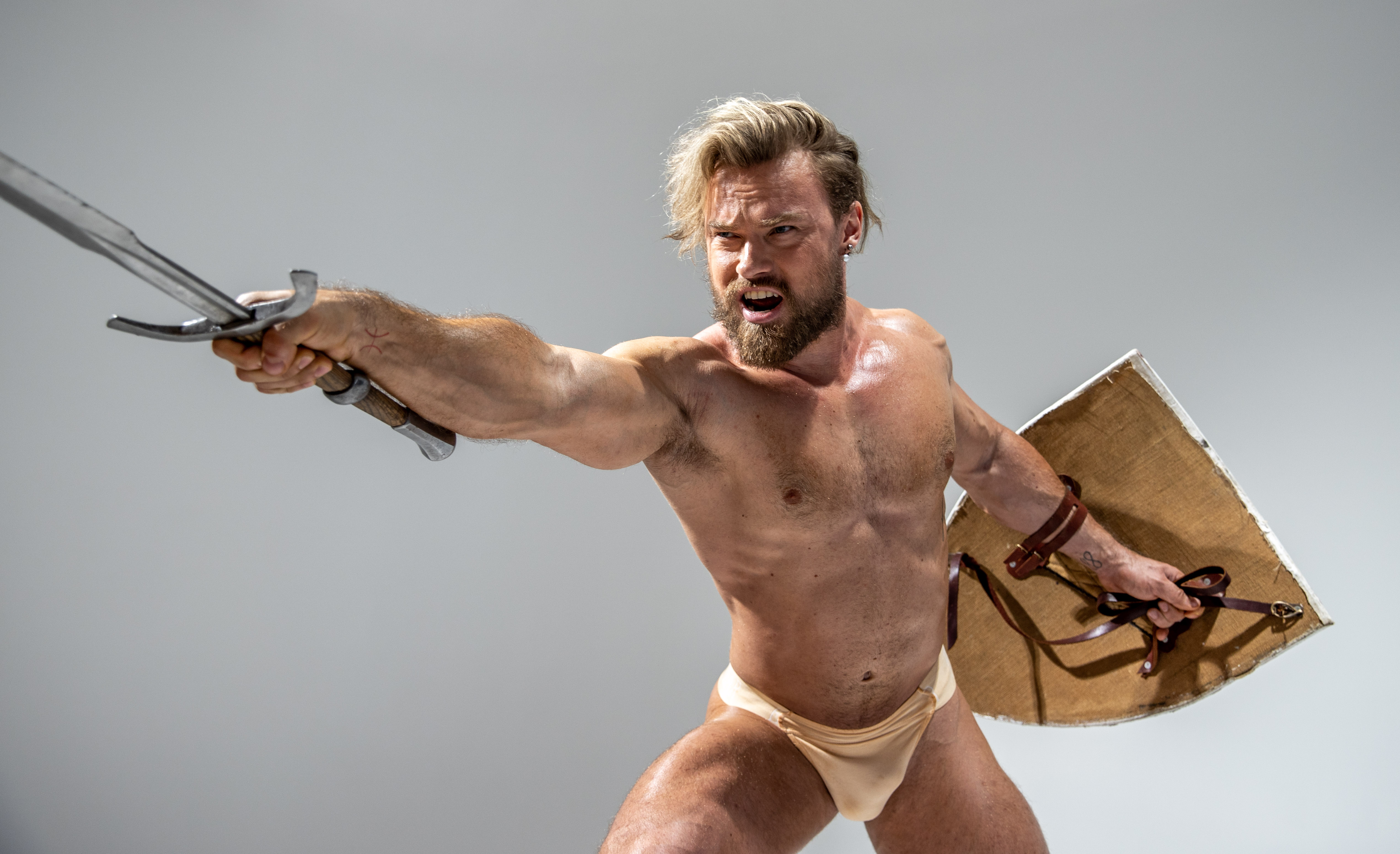 a muscular man with a beard and long blond hair, dressed only in beige briefs, strikes a dynamic pose against a light gray background. he brandishes a long silver sword in his right hand and holds a large round shield in his left. his expression is fierce and determined, as if engaged in battle. the image evokes themes of ancient warriors, mythology, and physical strength.