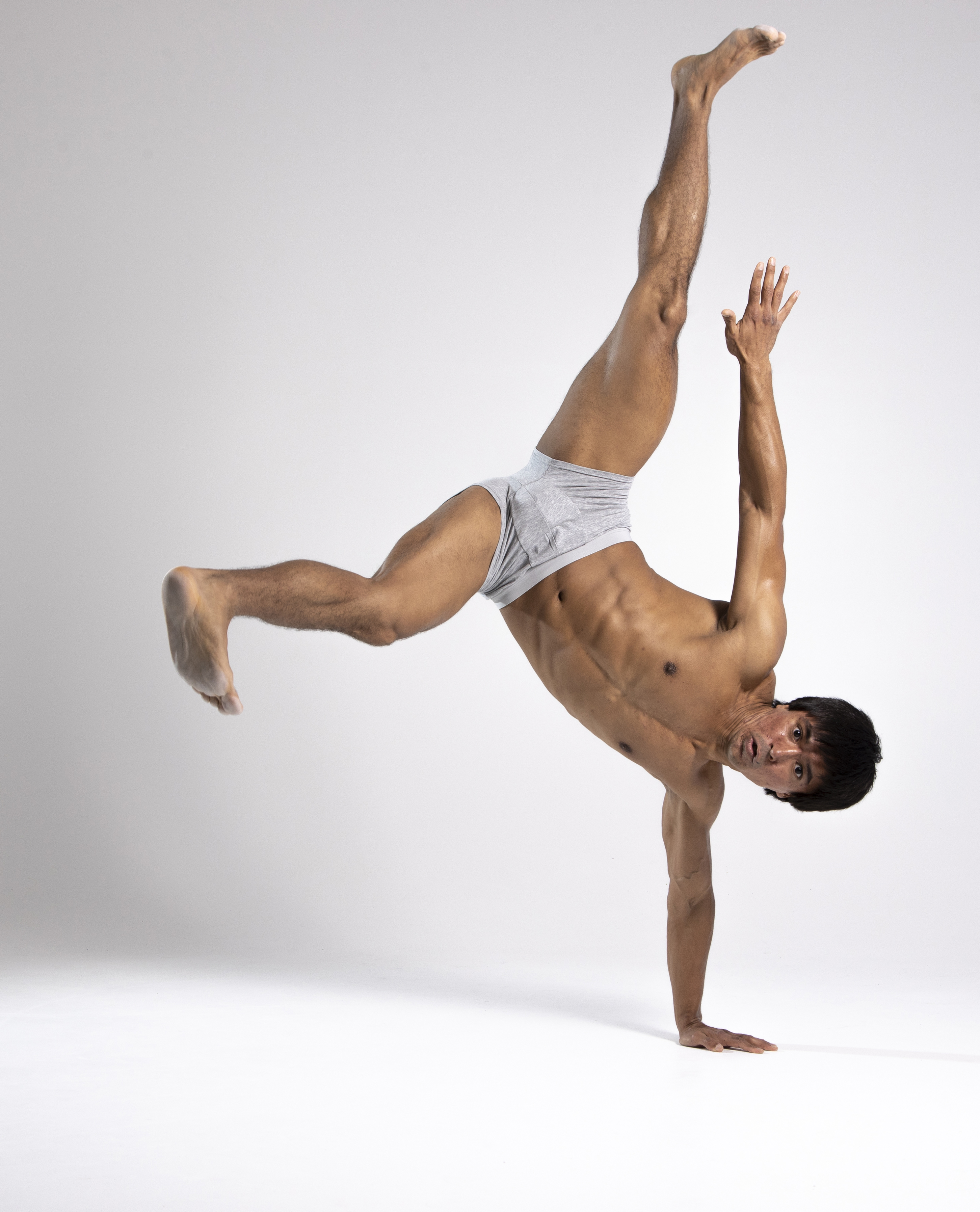 a muscular man in gray underwear performs a handstand in a studio setting. he is shirtless, showcasing his toned physique, and his body is angled, with one hand planted on the floor for support. one leg is extended upwards while the other is bent at the knee. the man is looking directly at the camera with a focused expression. the background is plain white, emphasizing the man's form and movements.