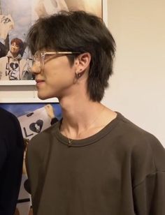 a side profile closeup shot of a man with glasses, featuring a short, shaggy haircut.  the man has tan skin and is wearing a brown shirt. the background is blurred and includes framed pictures featuring people.