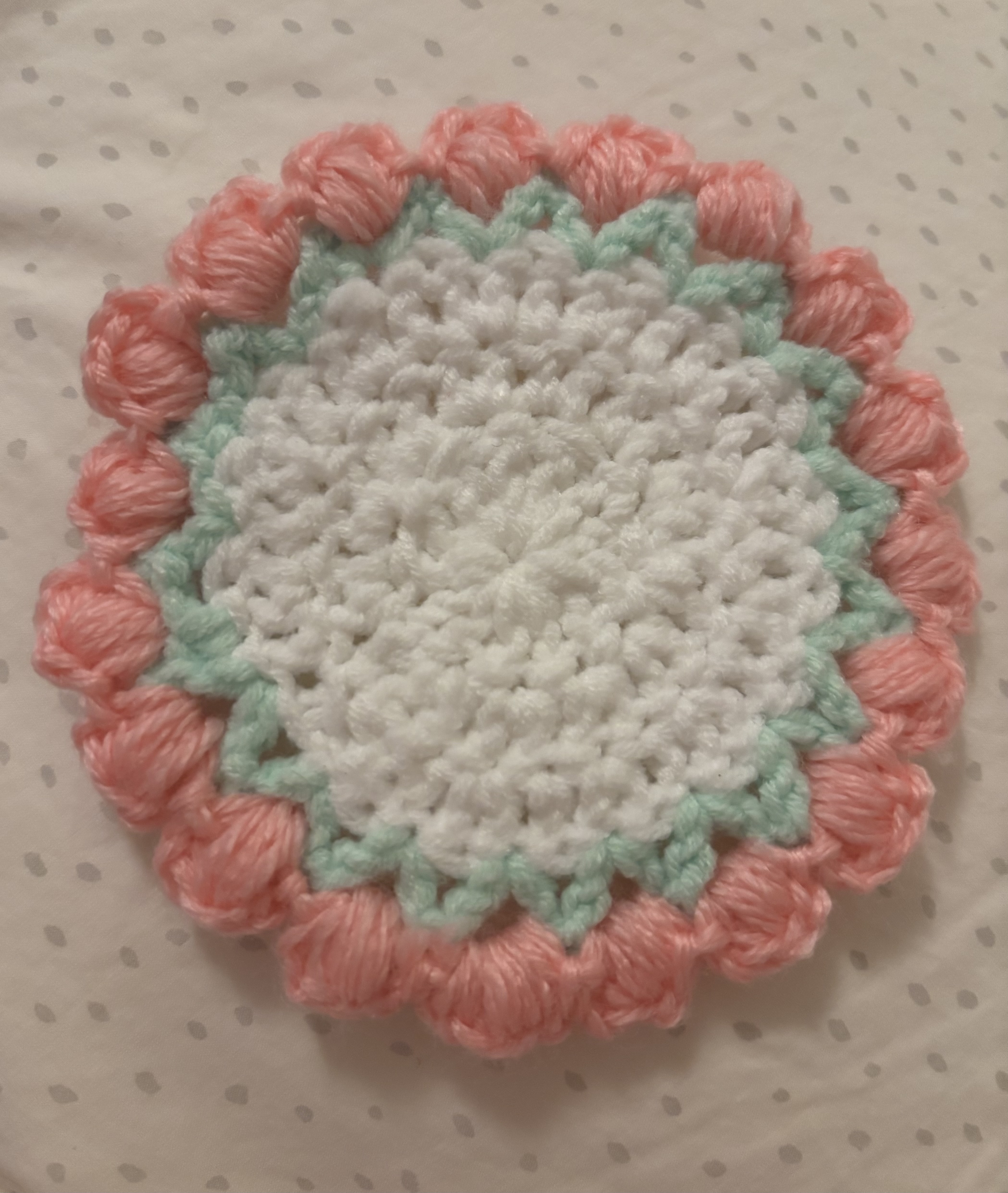 a closeup of a handmade crocheted doily. the doily is circular with a white center, a light blue border, and a pink outer edge with a bubbly texture. it is resting on a white surface with gray polka dots.