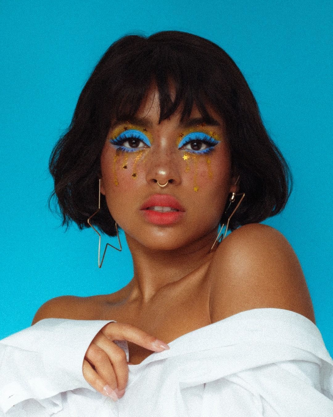 loseup portrait of a young woman with a bob haircut. she's wearing bright blue eyeshadow with gold starshaped glitter under her eyes.  she has freckles, a nose ring, and gold starshaped earrings.  her makeup is bold and colorful, and she's wearing a white offtheshoulder shirt. the background is a solid, vibrant blue. the overall aesthetic is glamorous and expressive.