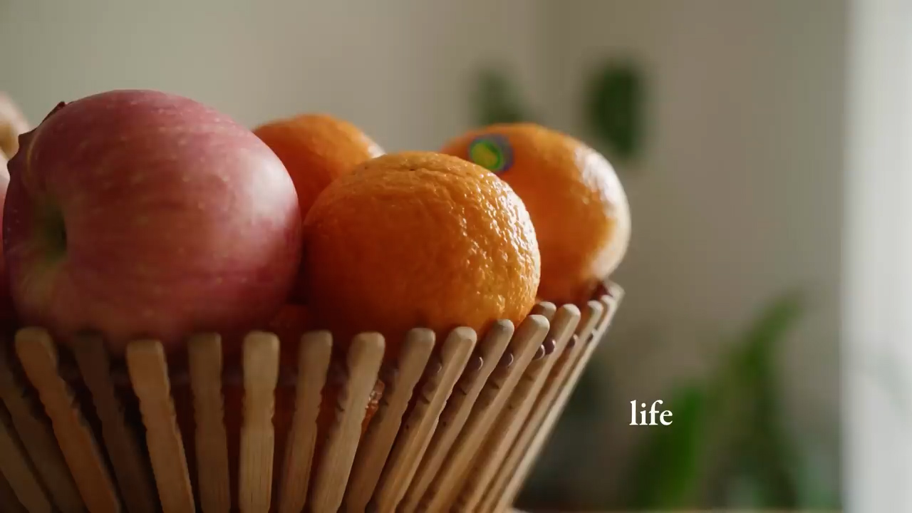 a wooden fruit basket filled with a red apple and three oranges sits on a table.  the background is blurred and out of focus.  the image gives off a natural, healthy vibe.