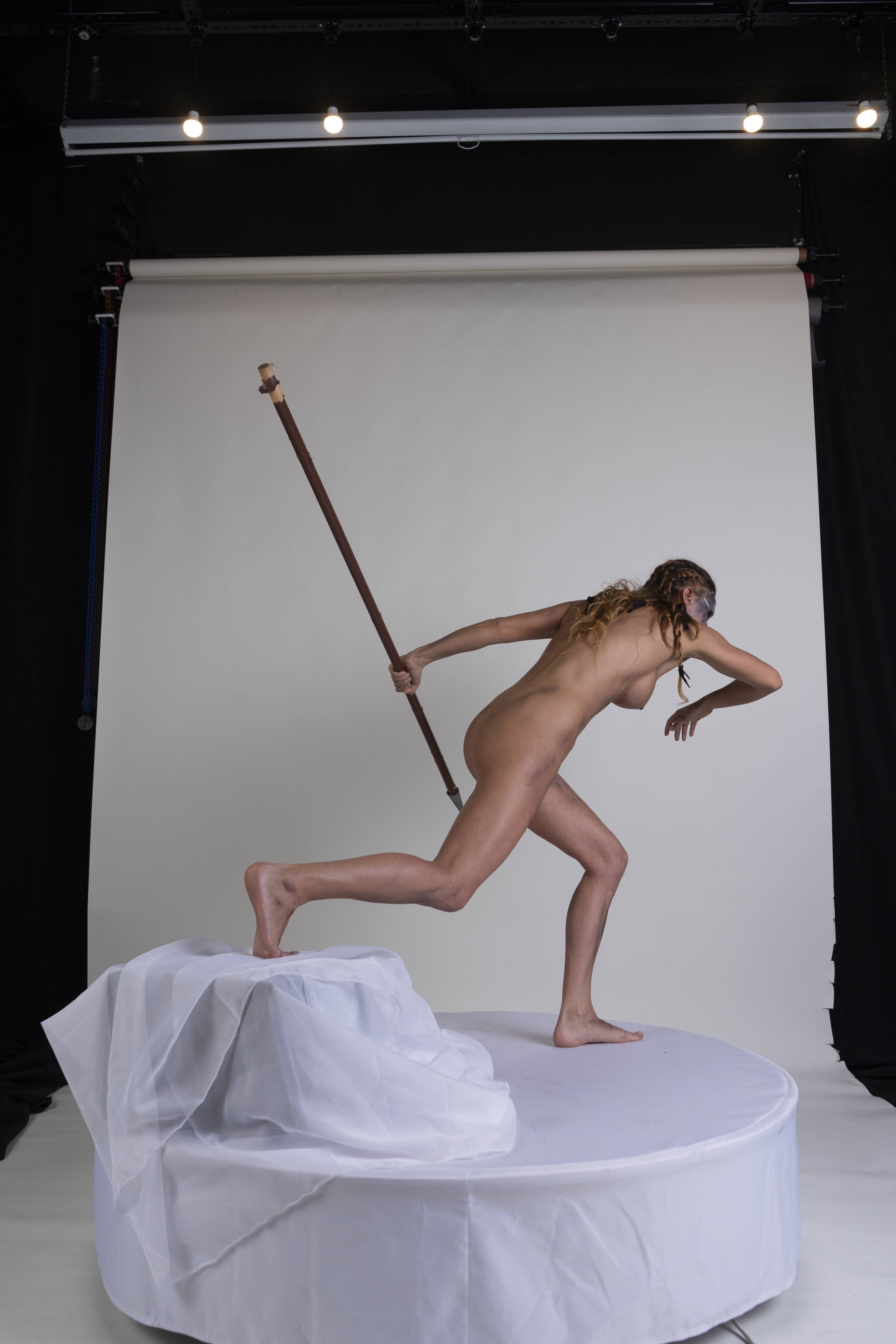 a nude woman stands on a draped platform, holding a spear. the background is a combination of black and white studio backdrops.