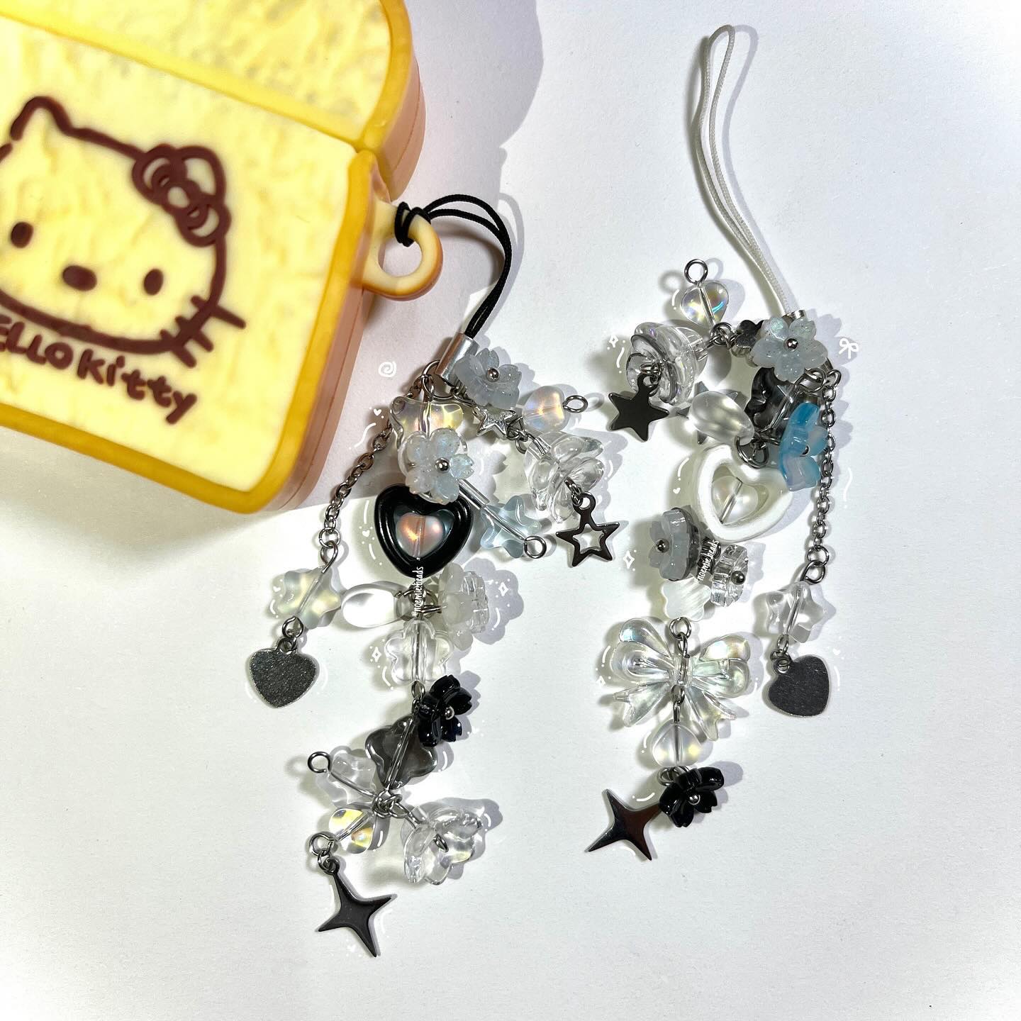 a closeup shot of two beaded earphone charms hanging next to a hello kitty case. the charms are made of various beads in white, clear, silver, and black colors, with heart, star, flower, and bowshaped beads dangling from chains. the hello kitty case is yellow and designed to resemble a piece of bread.