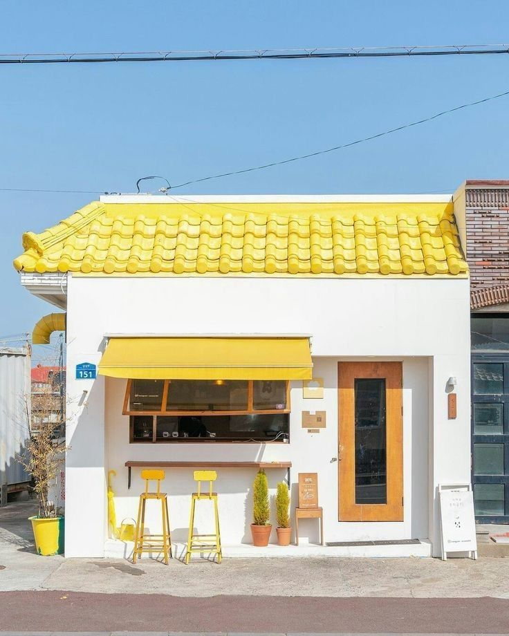 a small, charming cafe with a striking yellow roof and awning is the focal point. the building is primarily white with a wooden door and a large window shaded by the awning. two yellow stools are placed in front of the window, suggesting a seating area. potted plants add a touch of greenery, and various signs provide information about the cafe. the scene is set on a street, creating an inviting and picturesque atmosphere.