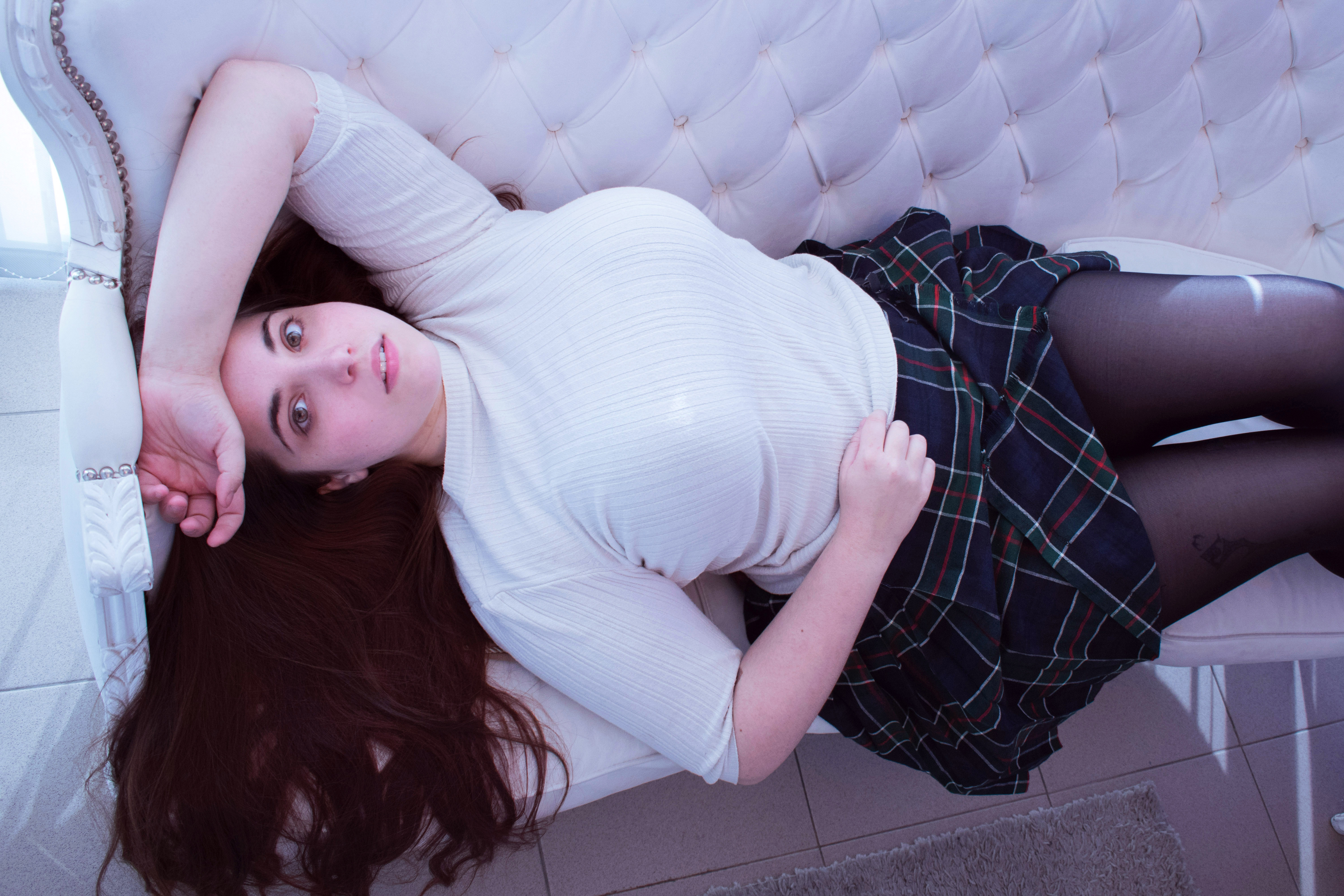 a woman with long auburn hair lies on a white couch, looking up at the camera. she is wearing a white, shortsleeved ribbed top, a dark blue and green tartan skirt, and black tights. her left arm is bent behind her head, resting on the couch's armrest. the couch has a tufted back, and a tiled floor is visible around it. the image is taken from a high angle.