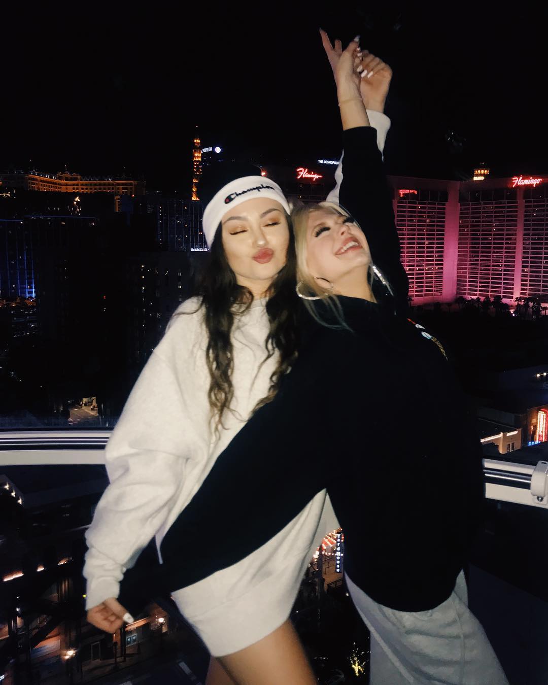 two young women posing for a photo at night with a cityscape in the background. the woman on the left has long brown hair and is wearing a white sweater and a black and white champion beanie. she is pouting her lips. the woman on the right has blonde hair and is wearing a black hoodie and grey sweatpants. she has her arm raised with her fingers in a peace sign. the flamingo hotel and other las vegas buildings are visible in the background. the woman on the right is also wearing large hoop earrings.