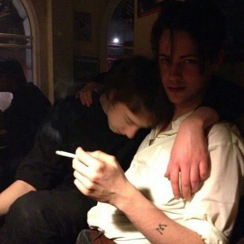 two people, seemingly men, are indoors. one man has his arm around the other and is holding a lit cigarette in his hand. a small tattoo is visible on his arm. the lighting is dim, creating a sense of darkness.