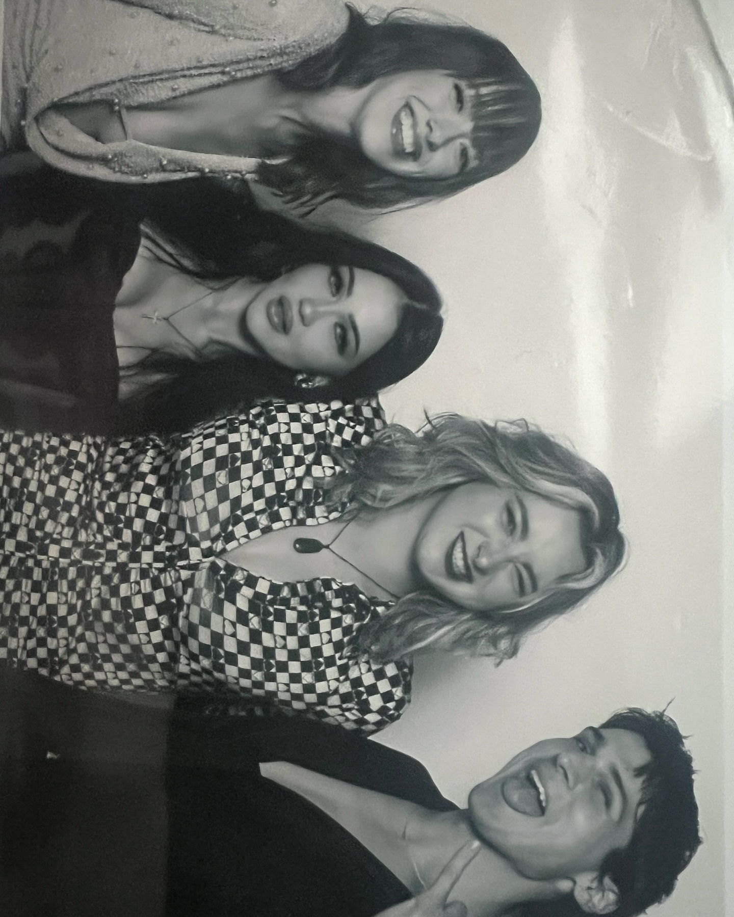 a black and white photograph shows a group of five young women and one young man posing together. the women are stacked on top of each other in a playful manner, smiling and looking directly at the camera. the woman on top is wearing a lightcolored jacket or sweater. the next woman is wearing a dark top, and the next has on a checkered shirt. the next woman has on a simple top, and the last is in dark clothing. the young man is at the bottom of the photo, grinning with his tongue out, arms around the other women.