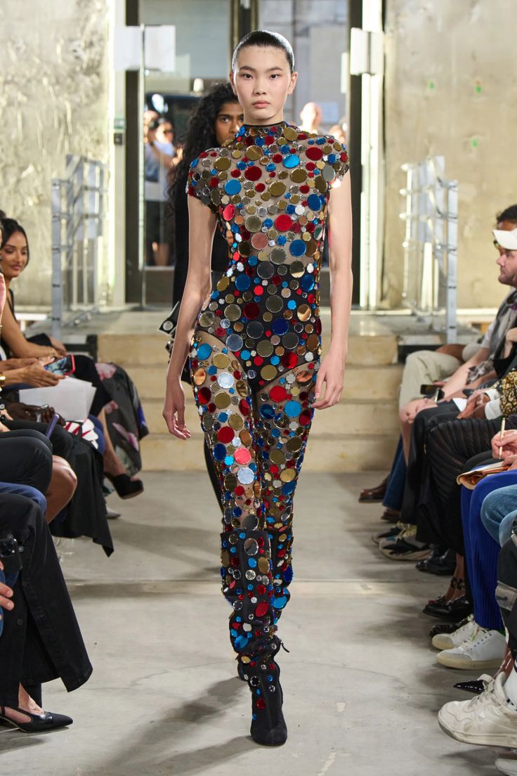 a fashion model walks down a runway wearing a unique jumpsuit. the jumpsuit is a sheer black fabric adorned with numerous circular mirrored pieces in various colors such as red, blue, and gold.  the model also sports matching high boots similarly decorated with mirrored pieces. the background shows a crowd of spectators seated on the floor, witnessing the fashion show. the setting appears to be a concretewalled venue. the overall aesthetic is discothemed, with a glamorous and stylish presentation.