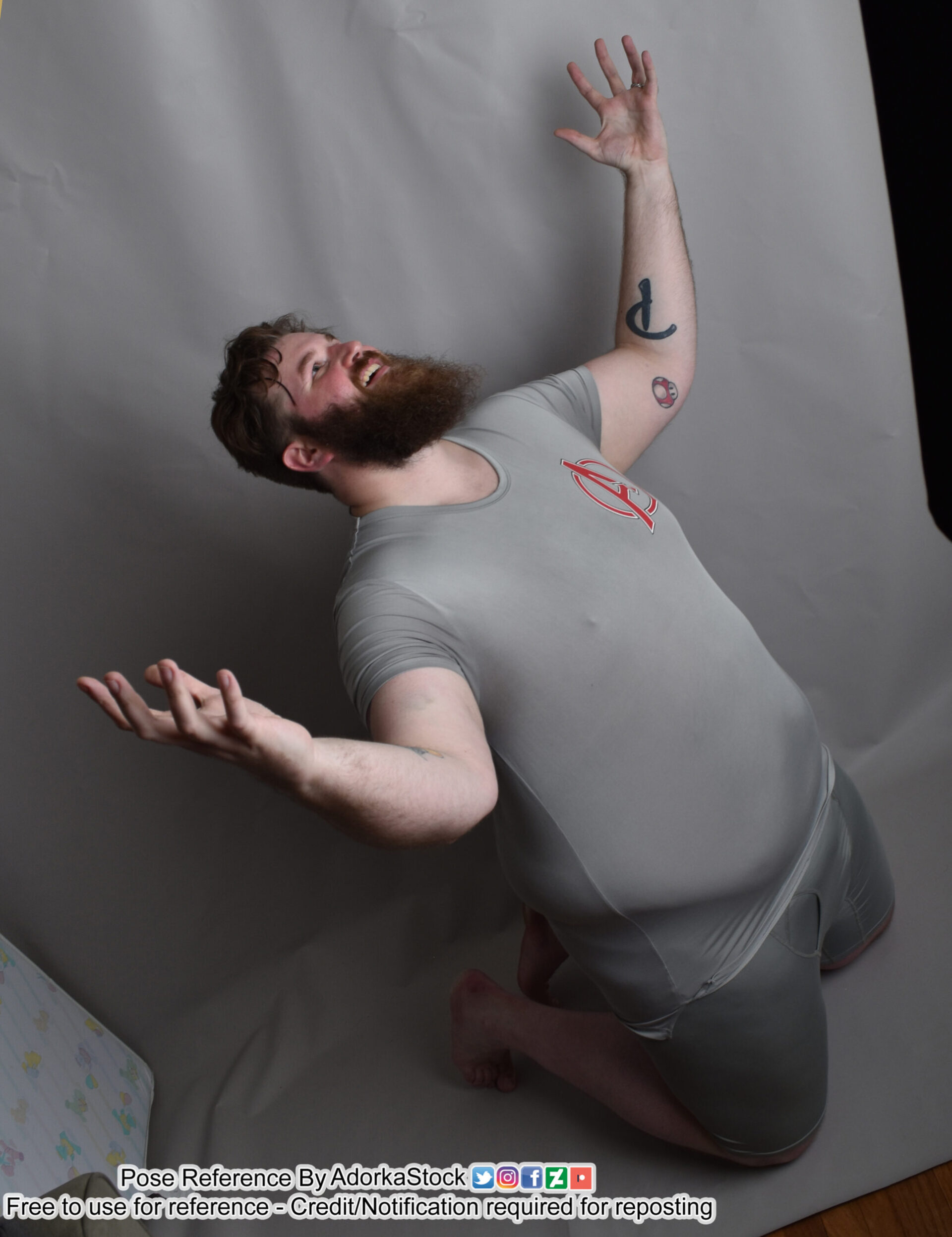a man in a grey shirt and shorts is kneeling with his right arm raised, reaching towards the camera. the image is a pose reference, with the man's body in a relaxed, natural pose. the image is taken in a studio setting against a grey backdrop.